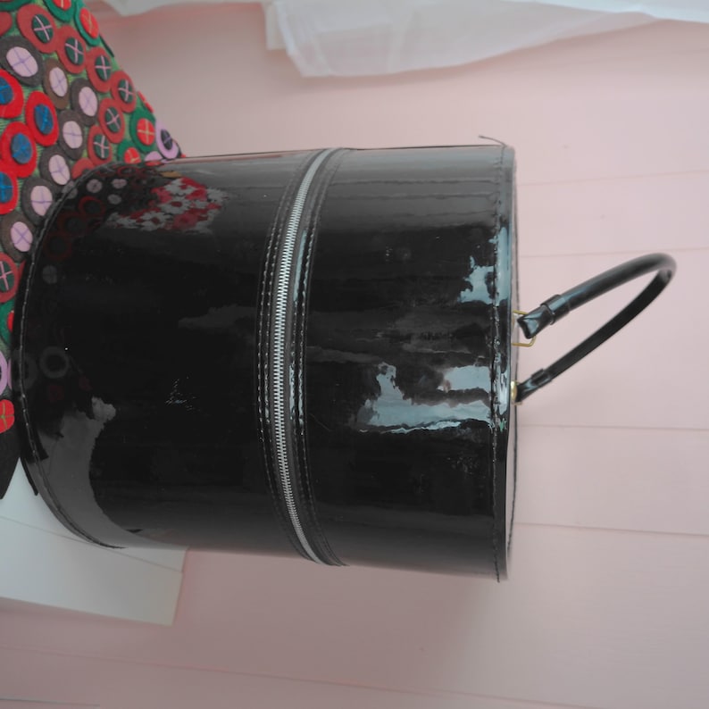 May include: A glossy black cylindrical handbag with a silver zipper running down the center. The bag has a curved handle and a shiny finish. The bag is a fashion accessory.
