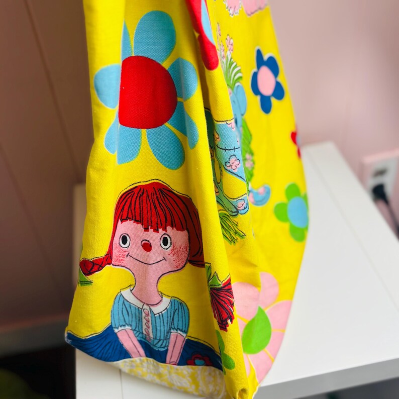 May include: A yellow fabric featuring a repeating pattern of large blue and red flowers, and a cartoon girl with red hair and a blue dress. The fabric has a vintage, retro aesthetic.
