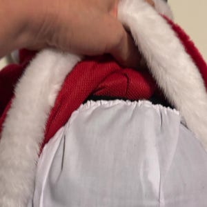 Pode incluir: Uma fantasia de Papai Noel vermelha com acabamento em pele sint&eacute;tica branca. A fantasia inclui um casaco vermelho com acabamento branco e forro branco. A imagem mostra um close-up da gola e do forro do casaco.