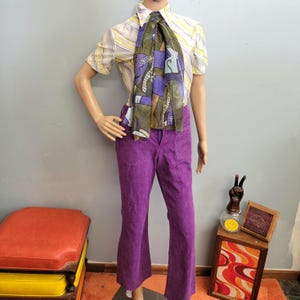 May include: A mannequin displays a vintage outfit. It includes a yellow and white striped short-sleeve shirt, a patterned scarf with purple and green accents, and bright purple corduroy pants. The outfit has a retro aesthetic.