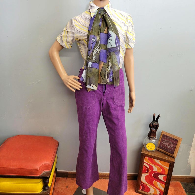 May include: A mannequin displays a vintage outfit. It includes a yellow and white striped short-sleeve shirt, a patterned scarf with purple and green accents, and bright purple corduroy pants. The outfit has a retro aesthetic.