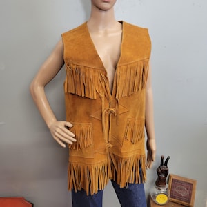 May include: A tan suede vest with fringe detailing. The sleeveless vest features a V-neck, two front pockets, and decorative star-shaped stitching. The vest has a tie closure and is approximately 24 inches long.