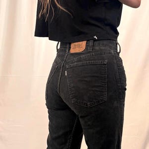 May include: A black short-sleeved crop top paired with black corduroy pants. The pants feature a tan leather-colored patch on the back waistband, a small white tab on the back pocket, and belt loops. The top has a small white logo on the hem.