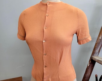 Puede incluir: Top de punto acanalado color melocotón con mangas cortas y cierre de botones. El top tiene cuello mandarín y costuras decorativas en los puños. La prenda se exhibe en un maniquí.