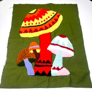 May include: A punch needle textile art piece on olive green fabric featuring three mushrooms. The central mushroom has a large orange, yellow, and red cap with geometric patterns. To the left is a brown and orange mushroom, and to the right is a white, blue, and pink mushroom.