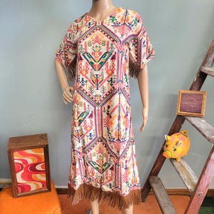 May include: A cream-colored midi dress featuring a vibrant, multi-colored geometric pattern with stylized figures and shapes in red, orange, green, and blue. The dress has short sleeves, a round neckline, and brown fringe trim along the sleeves and hem.