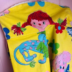 May include: A yellow fabric item with a whimsical design featuring a cartoon girl with red pigtails and a blue creature with pink flower accents. The fabric is decorated with colourful flowers and a pink dog-like character.