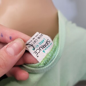 May include: Close-up of a light green t-shirt with a green neckline. A white tag is attached to the shirt, displaying the brand name "Spruce" and the size "Large 42-4". The tag also indicates the shirt is made of 100% cotton.