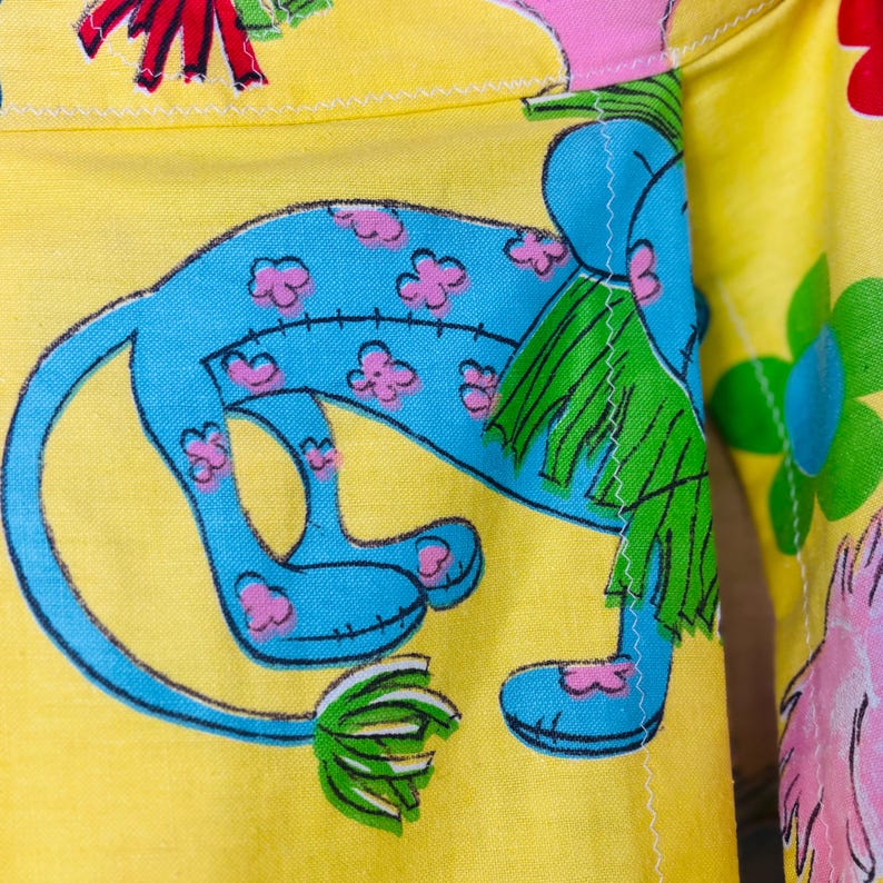 May include: Close-up of yellow fabric featuring a blue cartoon creature with pink flower details. The creature has green fringe and is set against a backdrop of colourful flowers. The fabric is stitched with white thread.