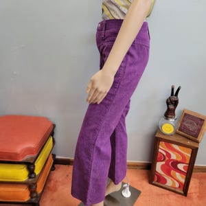 May include: A pair of purple corduroy pants with a flared leg. The pants are displayed on a mannequin. The mannequin is wearing a striped top. The pants have a classic five-pocket design.