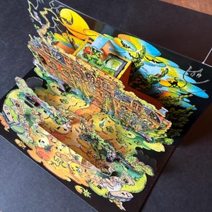 以下が含まれることがあります： 複数のレイヤーで構成されたお化け屋敷のシーンを描いたポップアップカード。カードは黒い背景に、お化け屋敷、木、満月のカラフルなイラストが描かれています。カードは開いており、3Dデザインが明らかになっています。
