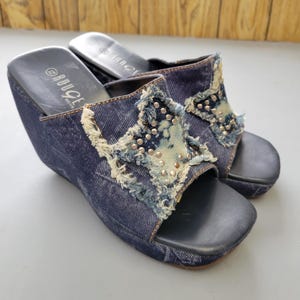 May include: A pair of denim wedge sandals. The open-toe shoes feature a dark blue denim material with a star-shaped design on the top, embellished with silver studs and frayed edges. The wedge heel is approximately 3 inches high.