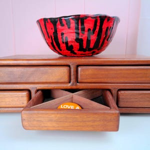 May include: A wooden desk organizer with five drawers, one of which is open to reveal an X-shaped divider and a small orange circular token with the text LOVE A. A ceramic bowl with a vibrant red and black drip pattern sits on top.