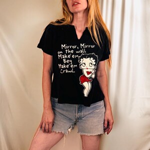 Könnte beinhalten: Ein schwarzes Kurzarm-T-Shirt mit Schlitz-Ausschnitt, das eine Betty Boop-Grafik und den Text Mirror, Mirror on the wall Make'em Beg Make'em Crawl zeigt. Dazu kombiniert ist eine hellblaue Jeansshorts mit ausgefranstem Saum.