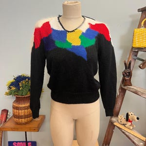 May include: A black sweater with a V-neck and long sleeves. The sweater features a colorful abstract design on the chest and shoulders, with patches of red, blue, green, and yellow. The shoulders have white and fluffy accents.