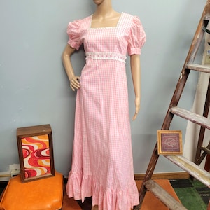 May include: A floor-length dress featuring a pink and white gingham pattern, short puff sleeves, and a square neckline. The bodice is accented with white lace trim along the neckline and waist, and the skirt finishes with a ruffled hemline.