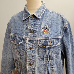 May include: Light blue denim jacket with a faded wash, featuring a Walt Disney Imagineering patch and Lee logo. The jacket has button closures, two chest pockets, and a classic collar. A vintage style jacket.