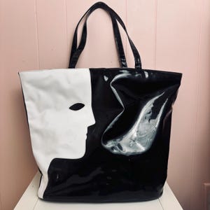 May include: A black and white tote bag with a glossy finish. The design features a face silhouette on the white side and a black background. The bag has two black handles for carrying.
