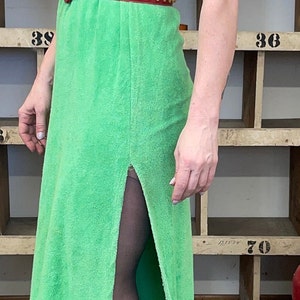 Puede incluir: Un vestido verde de felpa con capucha y un cintur&oacute;n rojo con tachuelas doradas. El vestido tiene una abertura lateral y la modelo lleva medias negras y sandalias de tac&oacute;n negras.