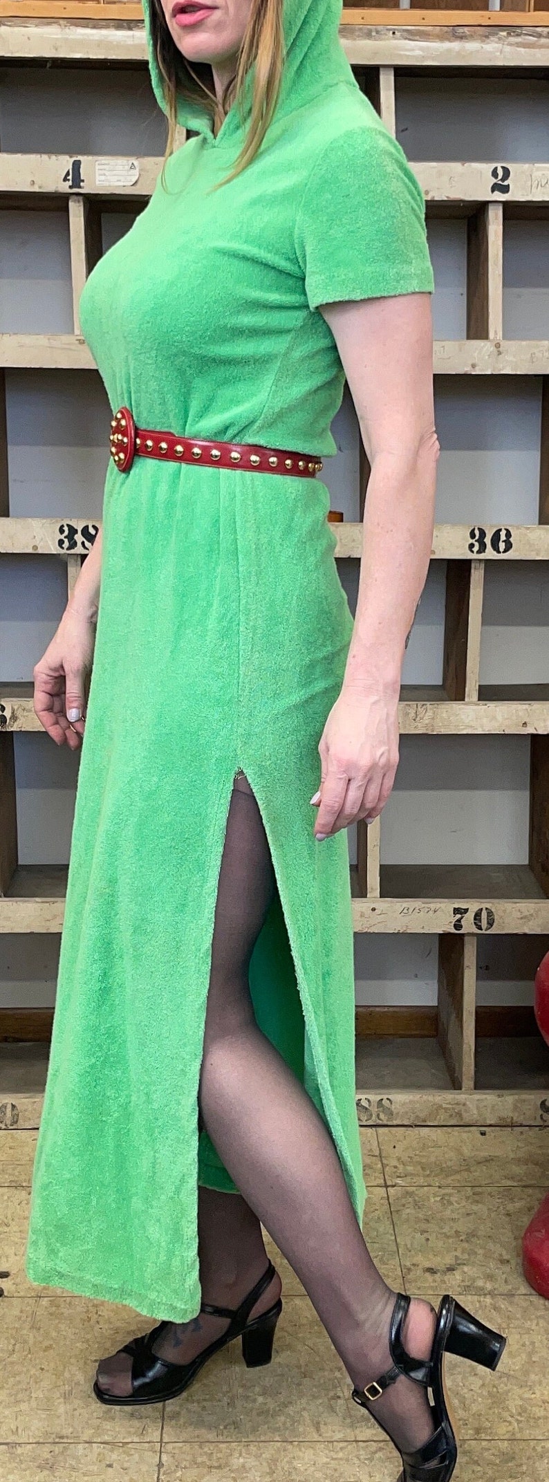 Puede incluir: Un vestido verde de felpa con capucha y un cintur&oacute;n rojo con tachuelas doradas. El vestido tiene una abertura lateral y la modelo lleva medias negras y sandalias de tac&oacute;n negras.