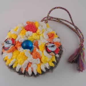 May include: A handmade, round, owl-shaped pendant with a colourful, textured crochet design. The owl features button eyes, a blue bead, and a tassel. The pendant is attached to a braided cord.