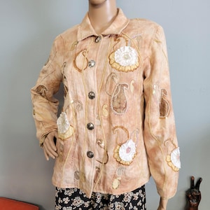 May include: A tan button-down jacket with floral and paisley embroidery. The jacket features decorative stitching, sequin accents, and large, dark buttons. White and yellow fabric flowers are sewn onto the front and sleeves. The jacket is displayed on a mannequin.