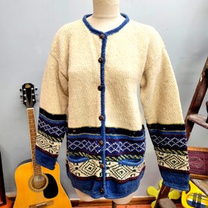May include: Cream-colored cardigan with blue trim and geometric patterns. The button-down sweater features a round neckline, two front pockets, and wooden buttons. The sleeves have blue and black striped cuffs with a geometric design.