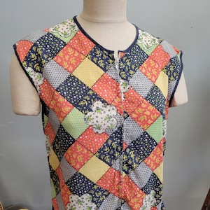 Może przedstawiać: Bezrękawnik pikowany z patchworkowym wzorem. Kamizelka posiada różnorodne kolorowe kwadraty w odcieniach czerwieni, żółci, niebieskiego, zieleni i bieli. Posiada zamek błyskawiczny na całej długości oraz granatowe wykończenie wokół dekoltu i otworów na ramiona.