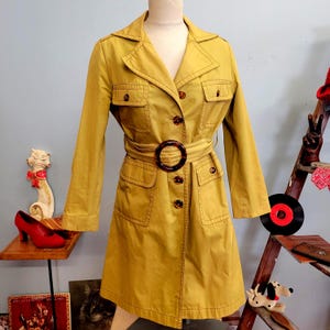 May include: A mustard yellow trench coat featuring a notched collar, long sleeves, and a belted waist with a large, round, tortoiseshell-patterned buckle. The coat includes two chest pockets and two hip pockets, all with flaps and tortoiseshell buttons.