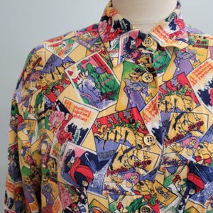 May include: A button-down shirt with a colorful print featuring illustrations and text. The shirt has a yellow background with various images in red, green, purple, and blue. Gold buttons accent the front.