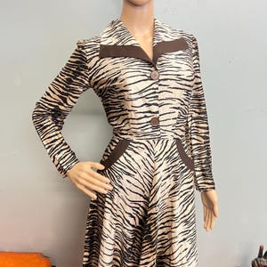 May include: A long-sleeved dress featuring a tan and black tiger stripe pattern. The garment includes a pointed collar with solid brown lapel accents, two brown buttons down the center front, and two brown pocket details at the waistline.