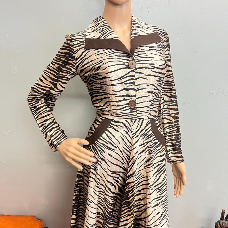 May include: A long-sleeved dress featuring a tan and black tiger stripe pattern. The garment includes a pointed collar with solid brown lapel accents, two brown buttons down the center front, and two brown pocket details at the waistline.
