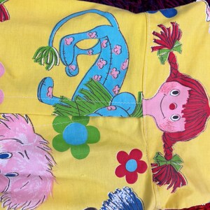 May include: Yellow fabric featuring a repeating pattern of cartoon characters and flowers. The characters include a blue creature with pink flower accents and a red-haired doll with pigtails. The flowers are blue, pink, and red.