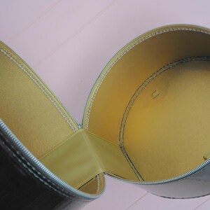 May include: Open, cylindrical, black case with a shiny finish. The interior is lined with a golden-yellow material. The case is open, revealing the interior. The zipper is silver.