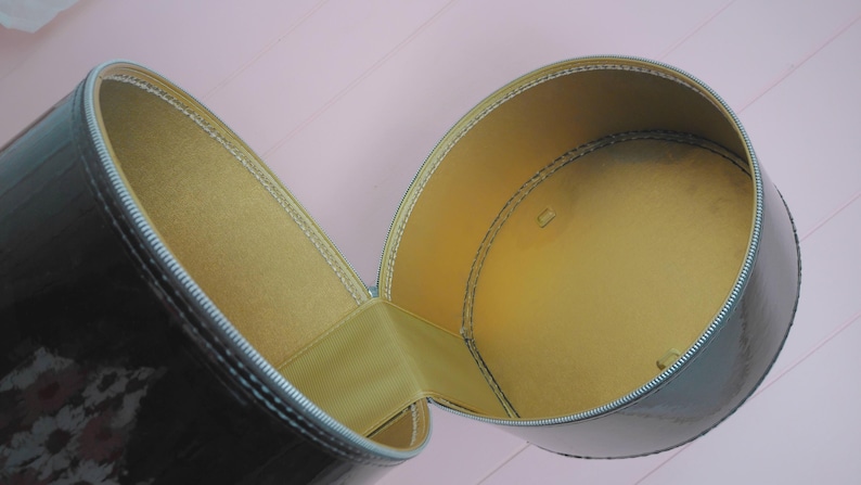 May include: Open, cylindrical, black case with a shiny finish. The interior is lined with a golden-yellow material. The case is open, revealing the interior. The zipper is silver.
