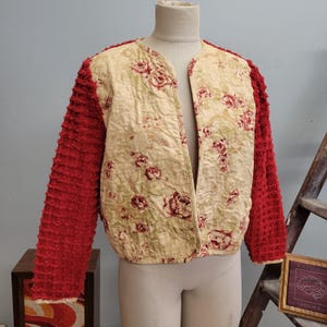 May include: A vintage jacket with a cream-colored floral pattern on the front and back panels. The sleeves are a vibrant red, textured fabric. The jacket is displayed on a mannequin, showcasing its open-front design and unique style.