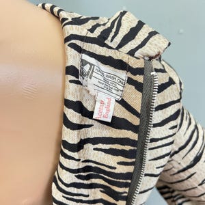May include: A garment featuring a black and beige zebra stripe pattern with a metal zipper closure. A white interior label reads HAND WASH ONLY, WARM (40 C), Hand-hot final rinse, Drip dry, and a second label reads Acetate, England.