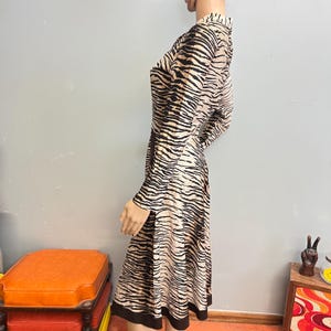 May include: A long-sleeved dress featuring a tan and black tiger stripe pattern throughout. The garment has a collared neckline, a fitted bodice, and a flared skirt that falls to the knees, finished with a solid black band at the hemline.