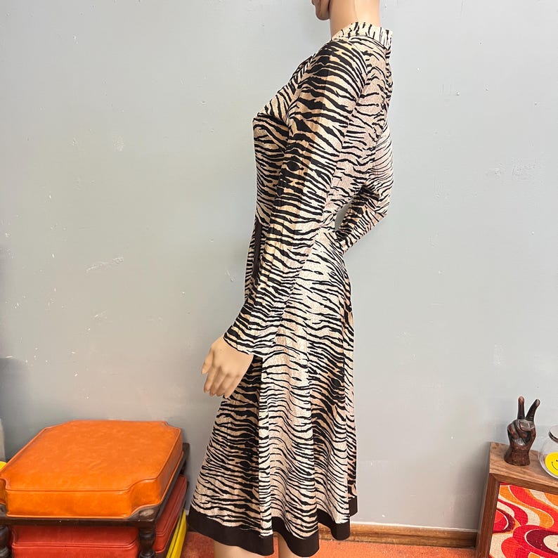 May include: A long-sleeved dress featuring a tan and black tiger stripe pattern throughout. The garment has a collared neckline, a fitted bodice, and a flared skirt that falls to the knees, finished with a solid black band at the hemline.