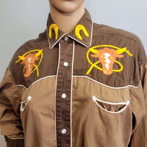 May include: Brown button-up shirt with embroidered bull and horseshoe designs. The shirt features a two-tone design with white trim and pocket details. The bull embroidery is orange and yellow.