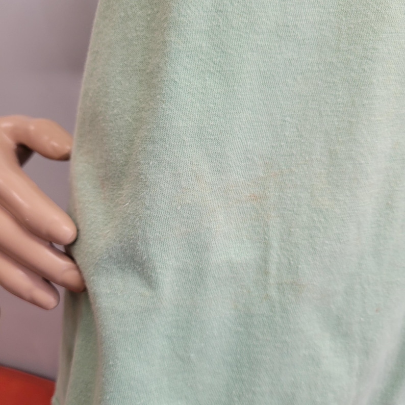 May include: A light green t-shirt with a slightly textured surface. The fabric shows some discoloration. The shirt is displayed on a mannequin.