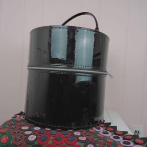 May include: A cylindrical black patent leather bag with a silver zipper and a curved handle. The bag is sitting on a colorful, patterned surface. The bag's glossy surface reflects the surrounding environment.