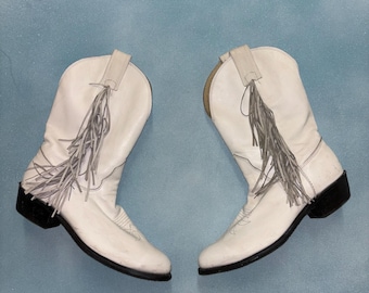 Peut inclure: Une paire de bottes de cowboy en cuir blanc avec des semelles et des talons noirs. Chaque botte est ornée de franges décoratives et d'une tirette en haut. Les bottes sont posées sur un fond bleu clair.
