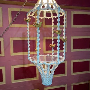 May include: A vintage macramé chandelier with a white woven design, featuring decorative gold-coloured bird accents. The chandelier hangs from a silver chain and has a basket-like bottom.