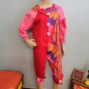 Puede incluir: Un colorido mono de payaso. El atuendo es principalmente rojo, con mangas y detalles rosas. Un lado presenta un estampado floral en naranja, morado y marrón. Pompones rosas adornan el frente y los puños son con volantes. La capucha está ribeteada con un volante.