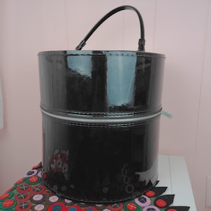 May include: A black, cylindrical makeup case with a glossy finish. The case has a silver zipper that goes around the middle and a black handle. The case is sitting on a colorful, patterned surface.