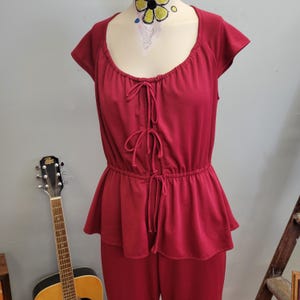 Puede incluir: Mono rojo con cuello redondo y mangas cortas. La parte superior presenta un detalle de cordón en la parte delantera y una cintura peplum. El mono se exhibe en un maniquí. Una guitarra es visible en el fondo.