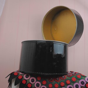 May include: A black, cylindrical hat box with a shiny, lacquered finish. The lid is open, revealing a gold interior. The box sits atop a decorative, colorful fabric with red, pink, and green accents.