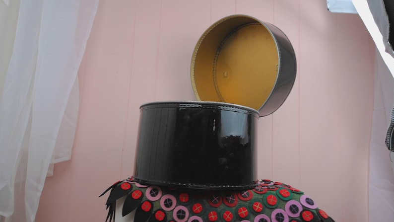 May include: A black, cylindrical hat box with a shiny, lacquered finish. The lid is open, revealing a gold interior. The box sits atop a decorative, colorful fabric with red, pink, and green accents.