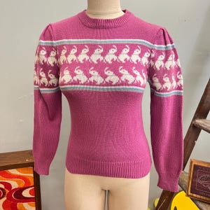 May include: A vintage magenta sweater with a crew neck and long sleeves. The sweater features a pattern of white elephants with light blue stripes across the chest and sleeves. The sweater is displayed on a mannequin.